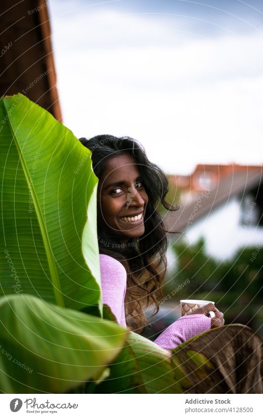 Smiling young ethnic female with cup of hot beverage on terrace woman positive balcony coffee morning rest warm smile drink beautiful cheerful relax fresh