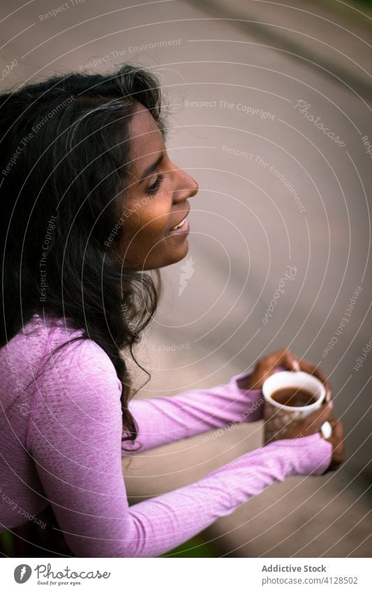 Smiling young ethnic lady with cup of hot beverage on terrace woman positive balcony coffee morning rest warm pink smile drink beautiful cheerful relax fresh