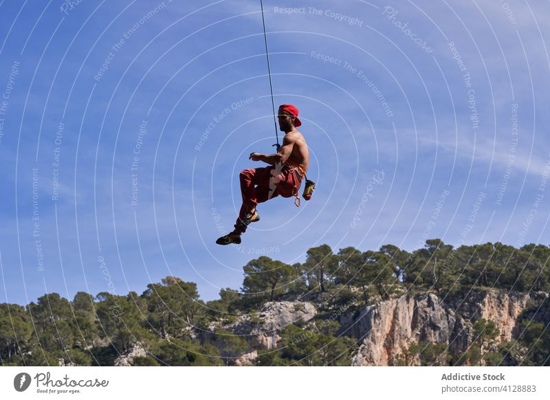 Strong male climber hanging on rope near rocky slope of mountain on sunny day man mountaineer strong sport secure equipment nature adventure activity cliff