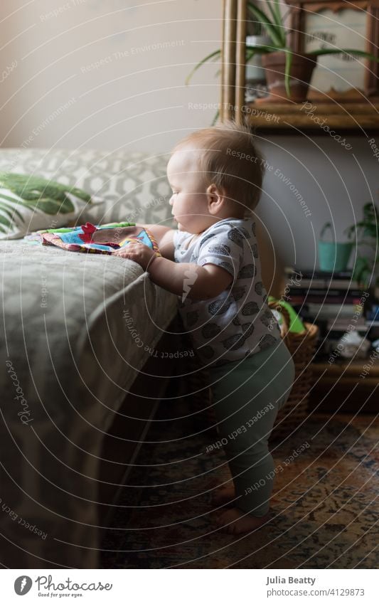 Young toddler standing up looking at a fabric children's book; eclectic decor with house plants infant 10 months old 6-12 months old baby young grasp manipulate