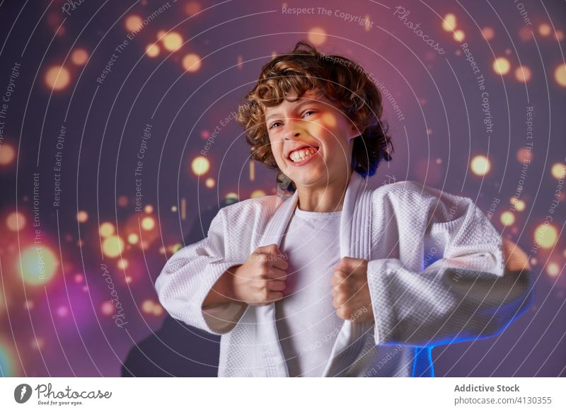 Excited boy in white kimono screaming among glowing lights power judo strong attack practice martial sport room expressive confident active loud happy smile