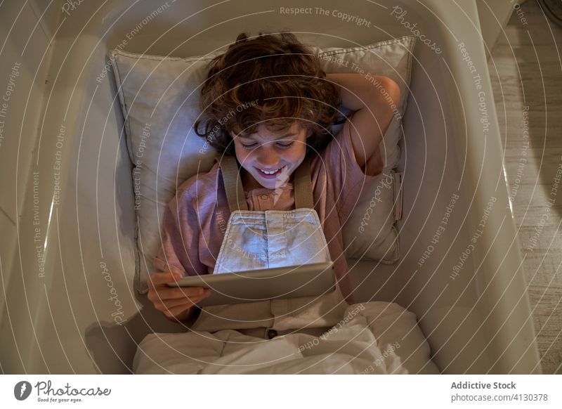 Cheerful boy using tablet in bathroom cartoon bathtub child cozy pillow fun lying watch weekend cheerful rest kid relax gadget device browsing curly hair