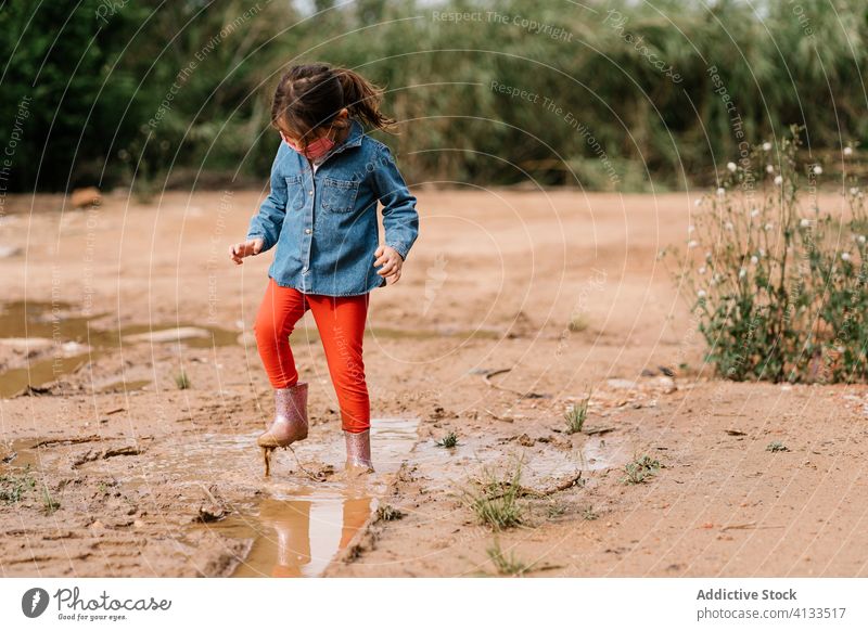 Curious girl surgical mask playing in mud puddle child having fun curious kid rubber boot cute dirty water wet adorable childhood joy activity playful ripple