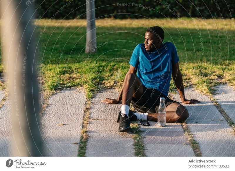 Athletic black man resting in the park african drinking water fitness sitting down strong flexing body muscle biceps arms athlete strength lifestyle athletic