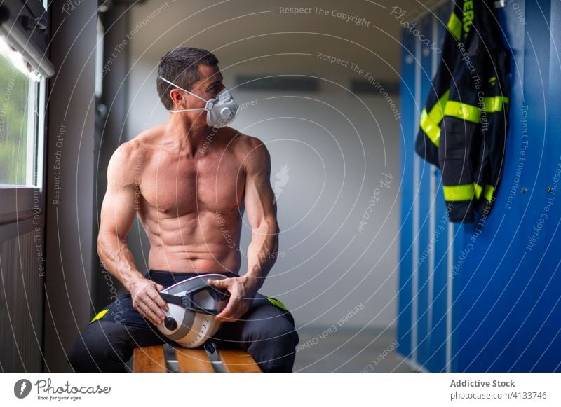 Muscular fireman in protective respirator at fire station firefighter safety work mature helmet male strong naked torso serious sit bench wooden muscular