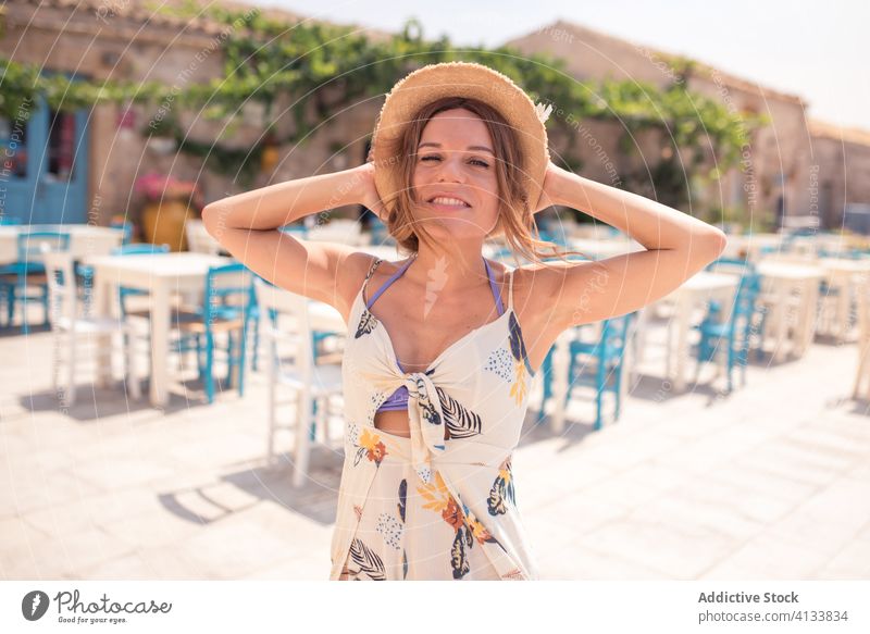 Stylish young woman in summer dress on cafe terrace in old town straw hat street exterior style building entrance plant aged allure facade outfit furniture