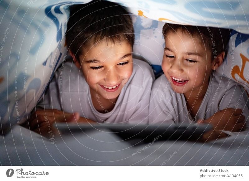 Happy brothers watching video on tablet together cartoon sibling browsing pajama blanket using hide enjoy home daytime interesting positive kid bed boy