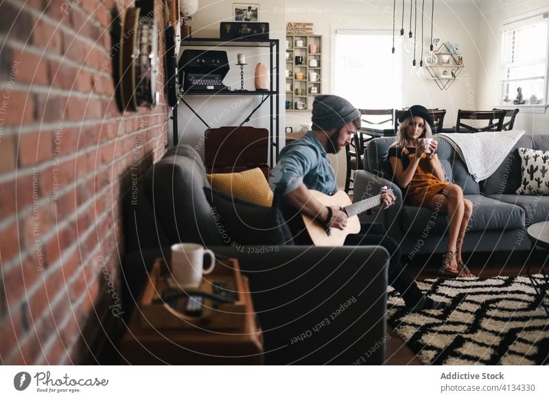 Musician playing guitar sitting with girlfriend on sofa at home couple music free time comfort living room friendship together relationship legs crossed