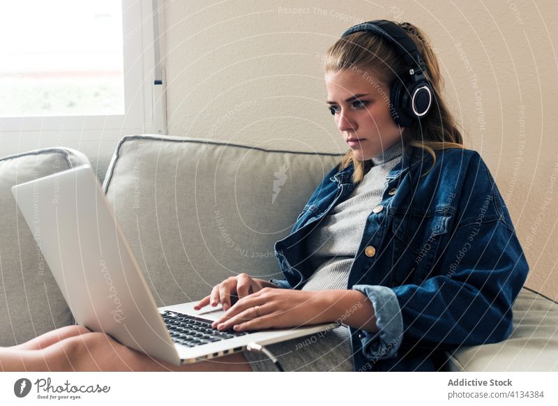 Modern woman working with laptop at home headphones sofa using focus concentrate young female casual wireless remote project freelance browsing device internet