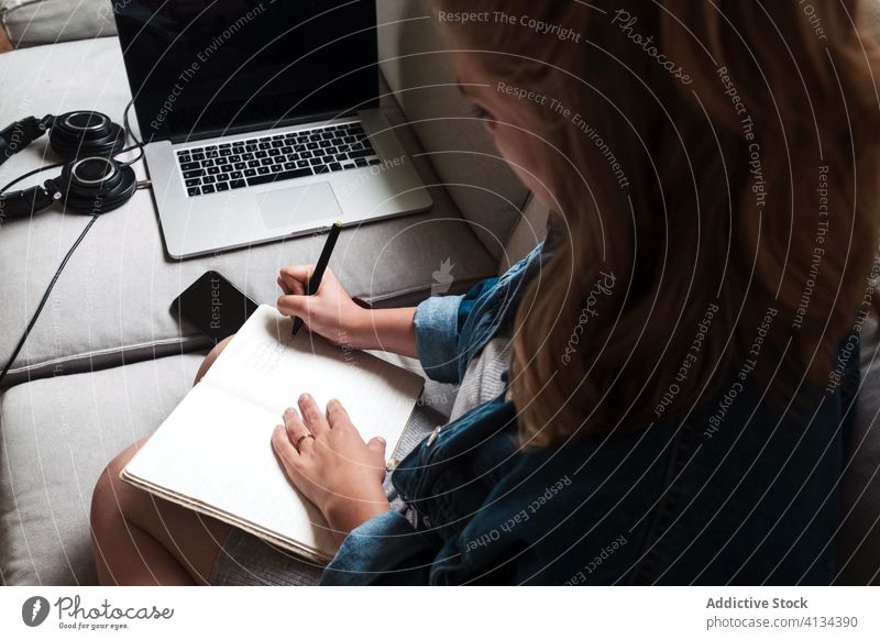 Millennial woman sitting on sofa with gadgets and writing in notebook write home young take note diary concentrate serious female casual focus device couch