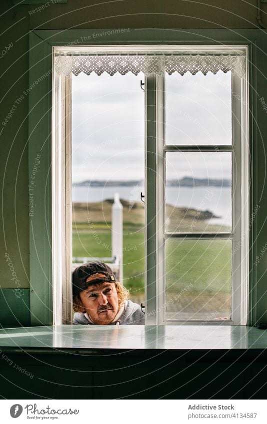 Mature man peering into window of house residential building open countryside rural curious male hipster mature adult cap rustic old fashioned cottage mustache