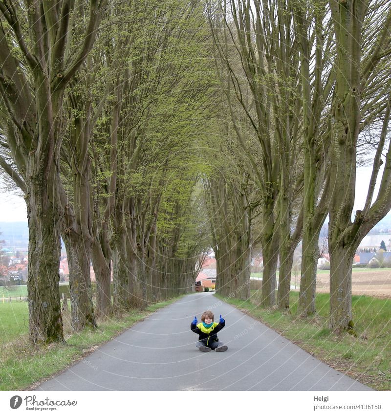 Child sits in the middle of the untraveled street of an avenue and gives the thumbs up Human being Schoolchild Street Avenue Asphalt Sit Spring Tree Field