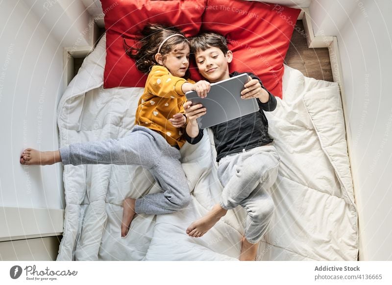 Friendly siblings using tablet together on blanket watch cartoon sister brother lying entertain relax girl boy weekend floor window lounge child kid childhood