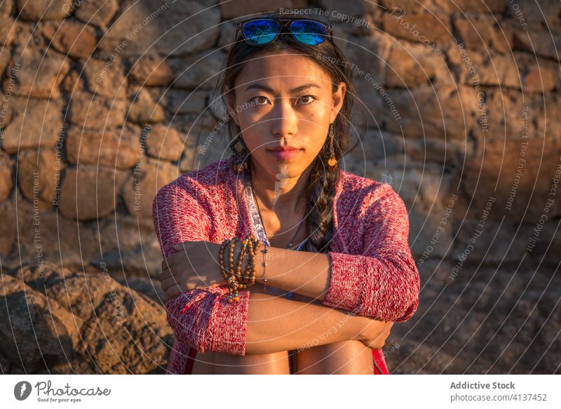 Young ethnic woman sitting against weathered stone wall young calm portrait ancient relax asian female style casual trendy accessory tranquil rest peaceful