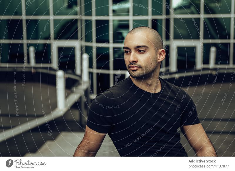 Positive ethnic man in trendy outfit sitting on street hipster young style positive building bald contemporary portrait modern guy casual ripped jeans beard