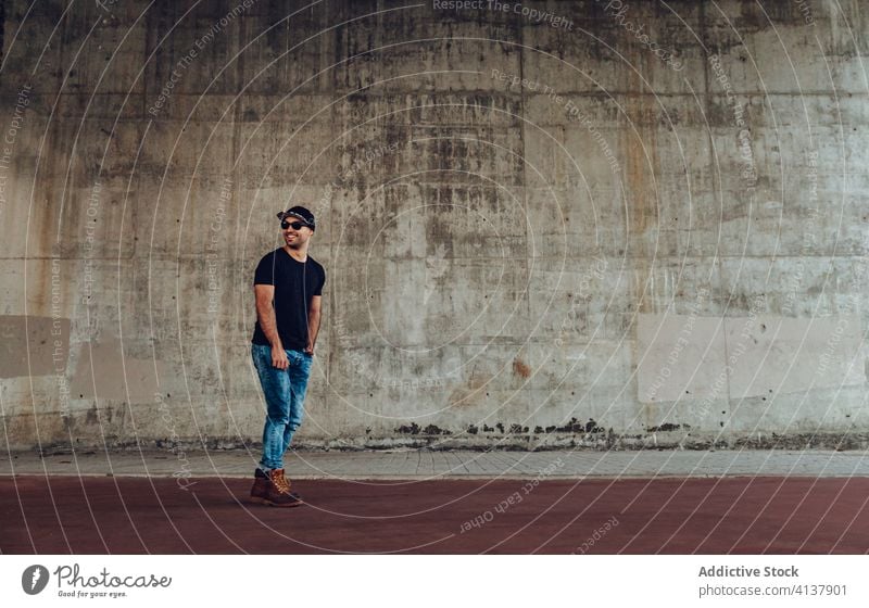 Young hipster man standing near building young style confident trendy modern wall concrete shabby happy male handsome guy casual beard outfit cloth jeans cap