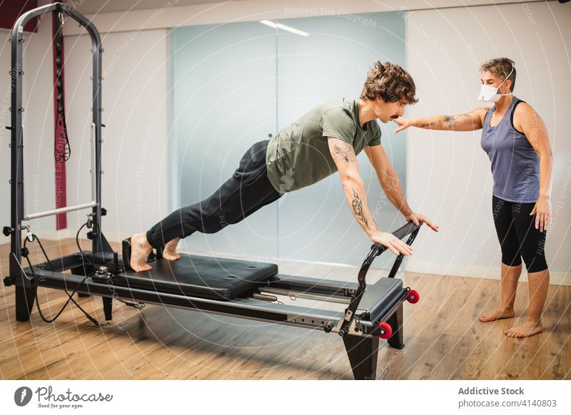 Sportsman using pilates machine under supervision of female trainer reformer sportsman exercise practice training together coronavirus gym respirator protect