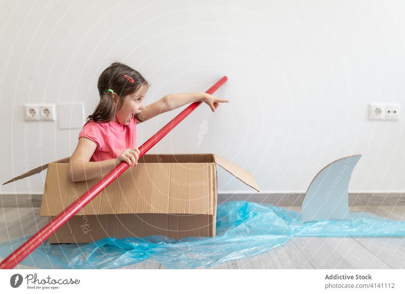 Joyful small girl in carton box sailing away from shark boat paddle game imagination fun playful kid sailor home child having fun joy pretend float childhood