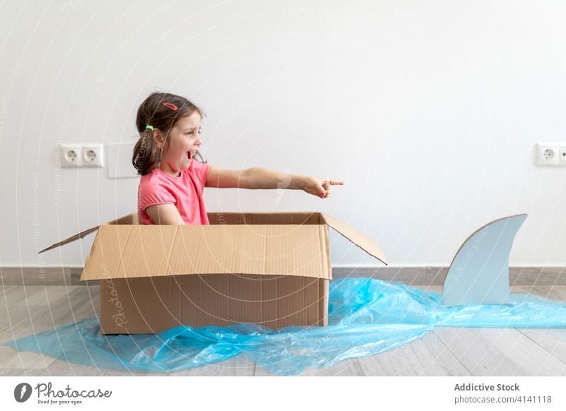 Joyful small girl in carton box sailing away from shark boat paddle game imagination fun playful kid sailor home child having fun joy pretend float childhood