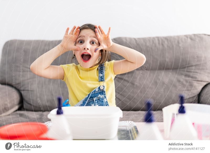 Expressive girl with dirty finger sitting on sofa at home grimace expressive mouth opened playful diy creative domestic child denim overall colorful amazed