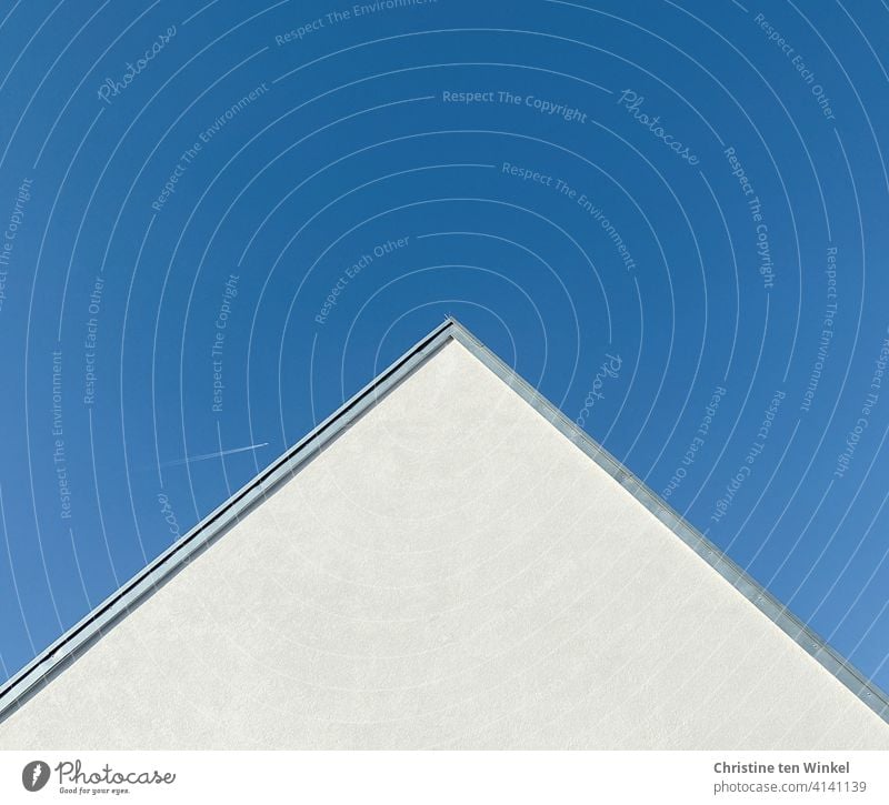 White plastered house gable with wires for bird repellent in front of bright blue sky. High up in the sky a tiny airplane with condensation trails. house gables