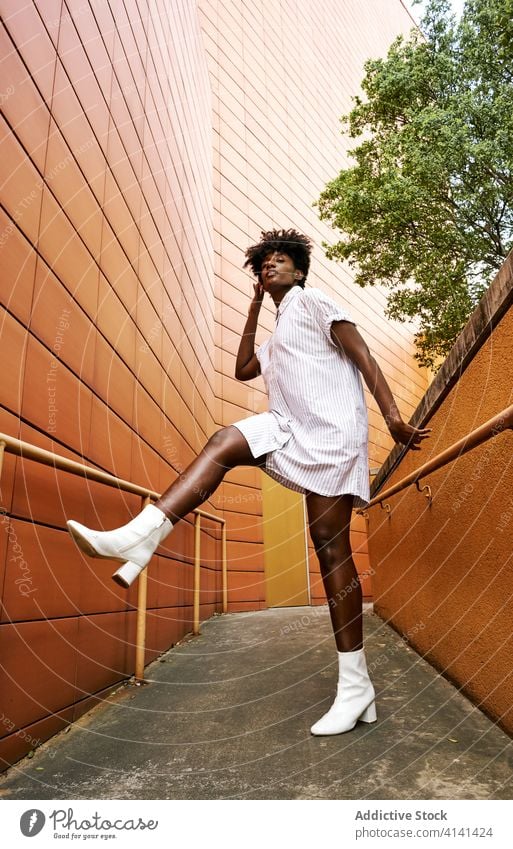 Confident African American woman in stylish outfit at entrance of contemporary building with orange tiled facade street fashion trendy exterior dress style