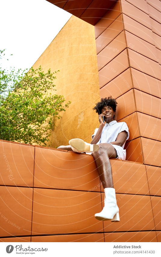 Stylish joyful black woman talking on phone on street near building with colorful exterior smartphone call architecture urban park chat modern conversation