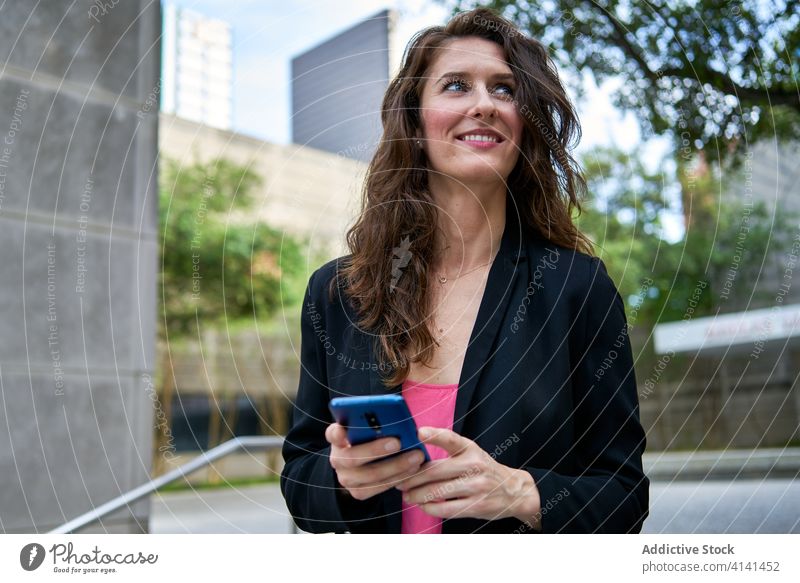 Cheerful businesswoman in jacket browsing smartphone on street entrepreneur using city classy smile urban female mobile device gadget communicate internet