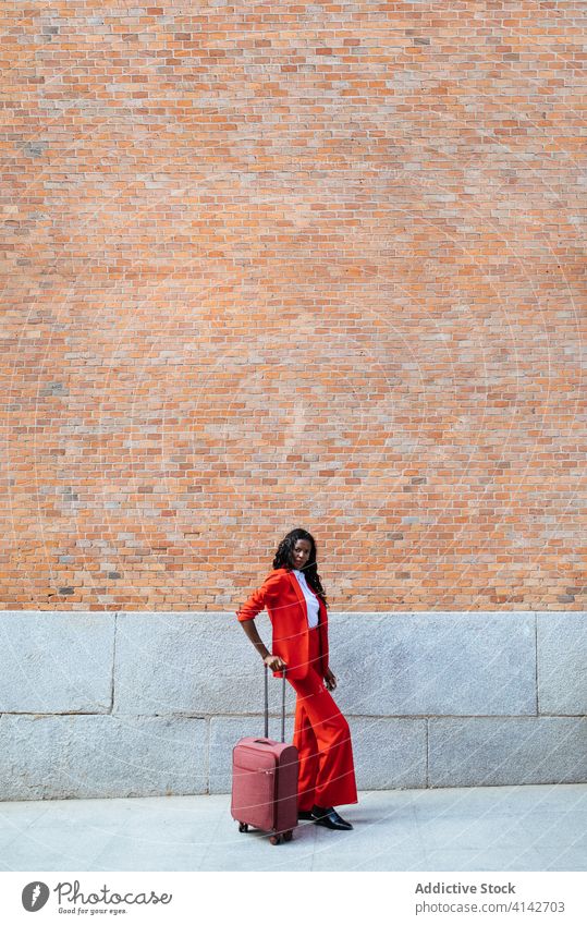 Black woman walking with suitcase on pavement fashion glamour style red brick wall building gesture elegant colorful appearance cloth stroll cheerful vogue