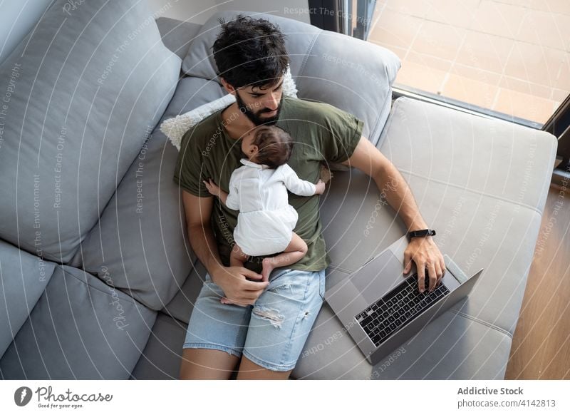 Busy father with infant using laptop baby work freelance sofa busy newborn project male fatherhood netbook home gadget remote browsing internet surfing online