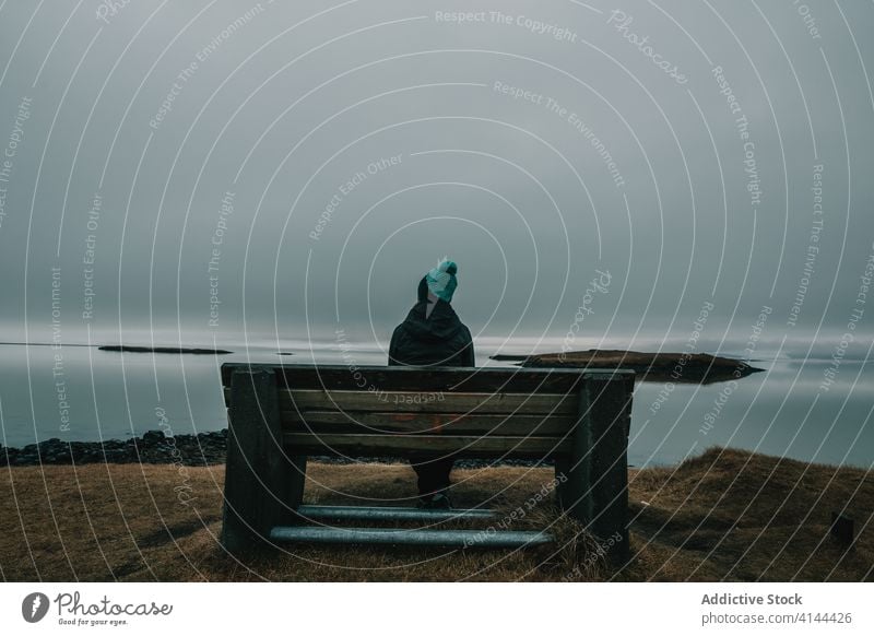 Unrecognizable traveler resting on bench near ocean against cloudy sky person relax hill admire tourist viewpoint overcast nature landscape iceland journey