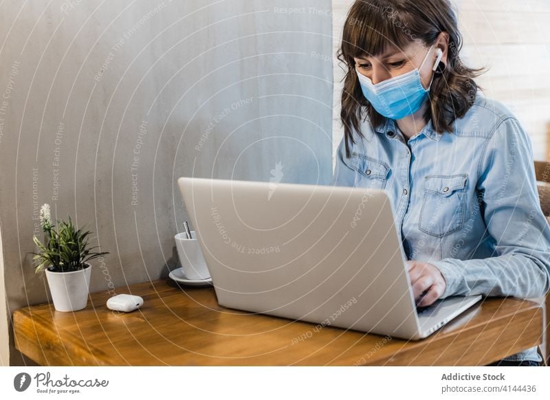 Concentrated woman working distantly on laptop in cafeteria freelance browsing earbuds focus medical mask coronavirus remote internet using female denim shirt