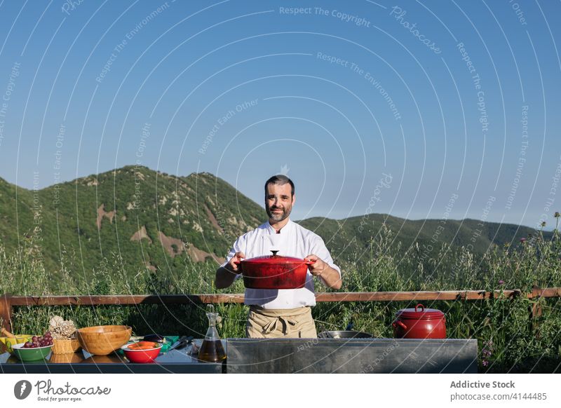 Chef in apron showing a pot in countryside man chef kitchen mountain terrain saucepan prepare professional job rustic restaurant cook uniform kitchenware work