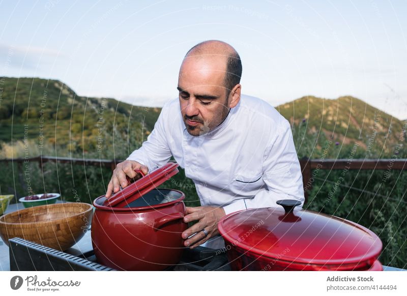 Focused cook checking dish boiling on stove in outdoor kitchen man smell saucepan dinner countryside prepare chef food professional meal male uniform