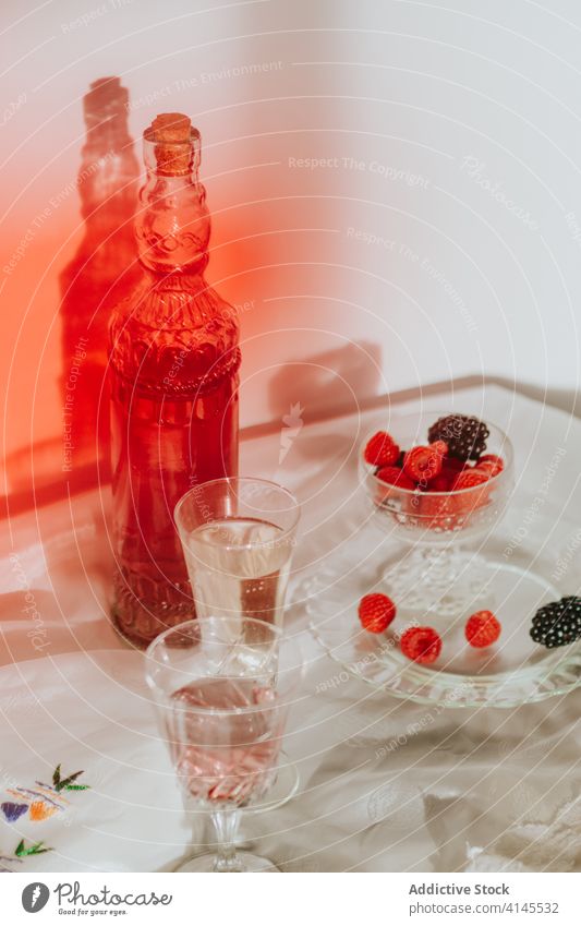 Glass of cocktail with berries and glasses served on table against white wall bottle plate berry drink composition decoration design creative elegant style