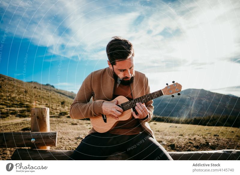 Bearded man playing ukulele in countryside fence sky cloudy music puerto de la morcuera spain male travel serious vacation trip nature rest journey sunny