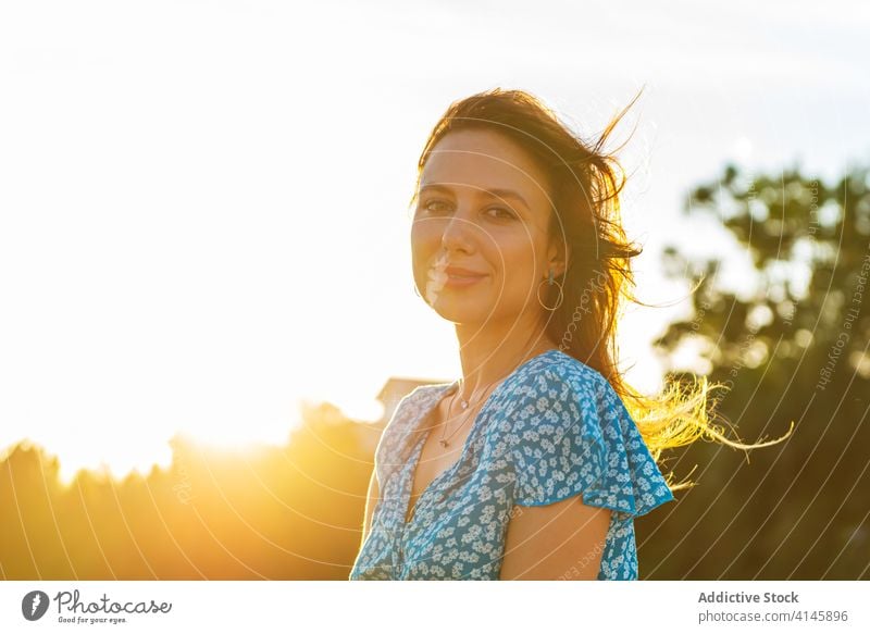 Happy young woman relaxing near green trees in countryside touch hair romantic wind sunset enjoy happy traveler smile cheerful rest female style trendy