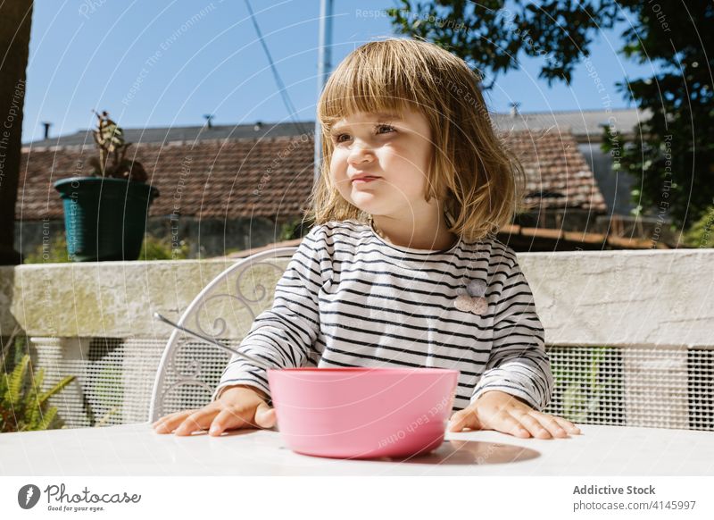 Charming girl sitting at table during breakfast adorable child morning terrace smile food bowl kid happy relax sunny summer courtyard backyard little healthy