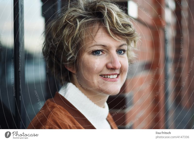 SMILE - EVENING SUN - BRICK Woman Curl Short-haired Short haircut wisps Blonde Smiling Brick Colour photo Adults Exterior shot Day Contentment Wait Anticipation