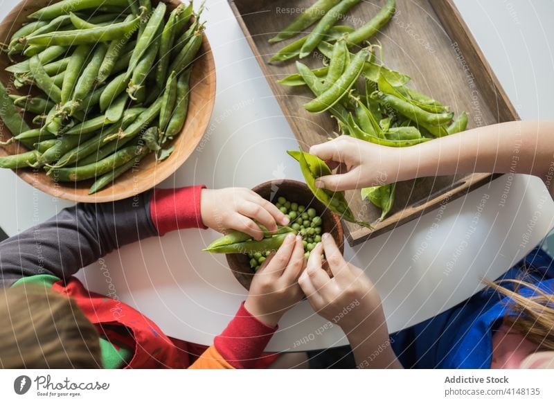 Crop children peeling pile of pea pods at home help fresh vegetarian bowl together sibling raw childhood uncooked product wooden round table green crunchy