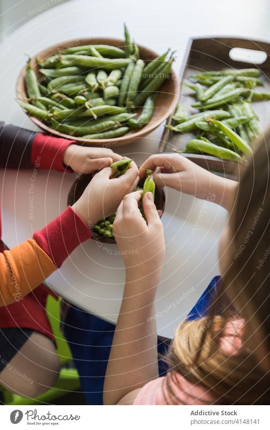 Crop children peeling pile of pea pods at home help fresh vegetarian bowl together sibling raw childhood uncooked product wooden round table green crunchy