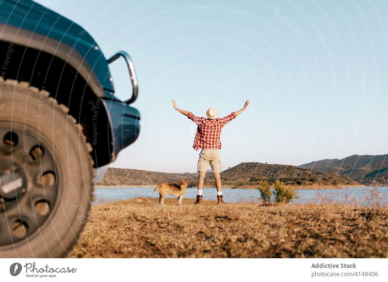 Unrecognizable traveler with dog contemplating river near mountains in summer nature contemplate hands apart tourism car lake freedom grass faded man trip