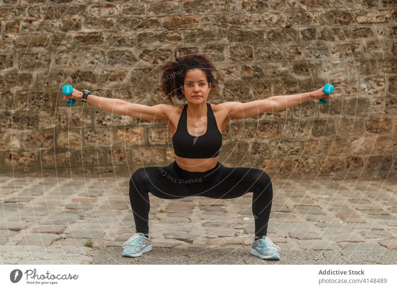 Fit ethnic sportswoman squatting with dumbbells on embankment during workout training balance reached arm pavement stretch practice flexible activewear strong