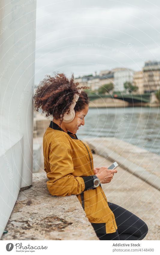 Sportswoman in headset using social media on smartphone after workout sportswoman headphones chatting internet break embankment river gadget device online