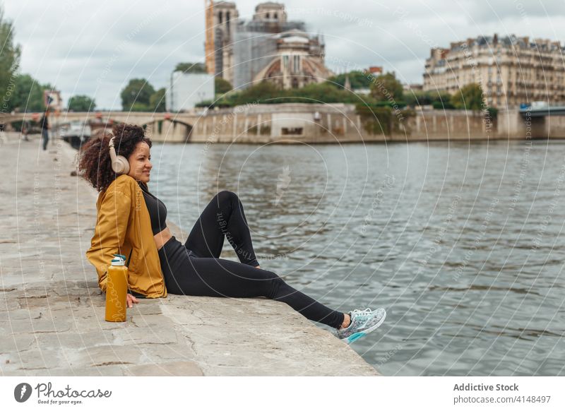 Ethnic sportswoman in wireless headset contemplating river during break contemplate lake listen music enjoy city smile water ripple sportswear using device