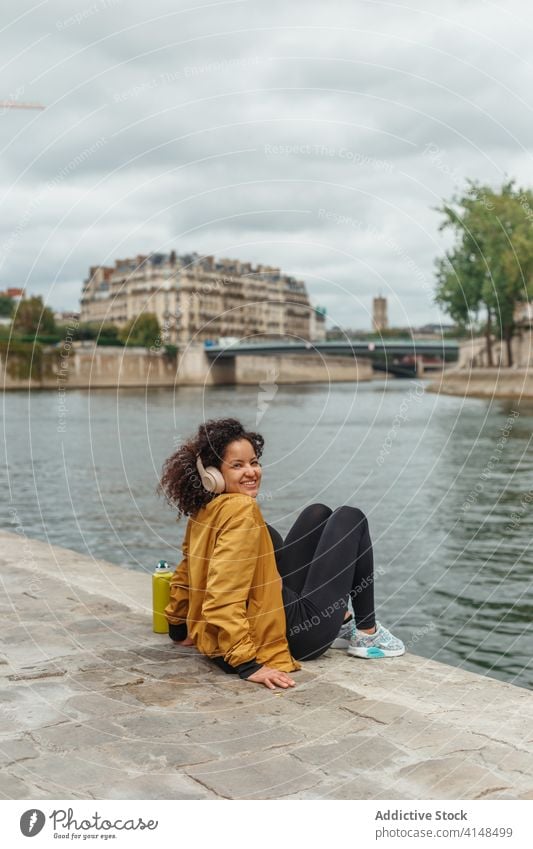 Ethnic sportswoman in wireless headset contemplating river during break contemplate lake listen music enjoy city smile water ripple sportswear using device