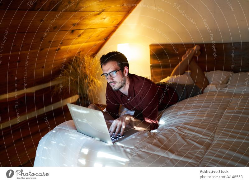 Concentrated male freelancer typing on laptop at home remote man work project entrepreneur using bed lying thoughtful browsing netbook internet device gadget