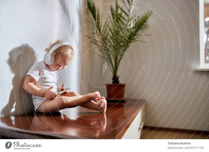 A child with headphones looks into a smartphone. The bright Scandinavian-style interior features a potted date palm. The rays of the morning sun in the room. Technology and people concept music at home. Copy space.