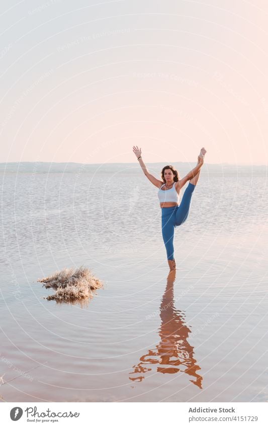 Slim woman stretching in yoga pose on lake shore practice asana stand split thiruvikramasana balance water calm harmony wellness lifestyle female flexible hatha