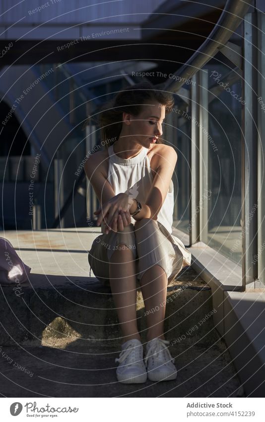 Relaxed young woman sitting near glass fence in city pensive relax style urban building thoughtful calm dreamy female modern fashion trendy street solitude lady